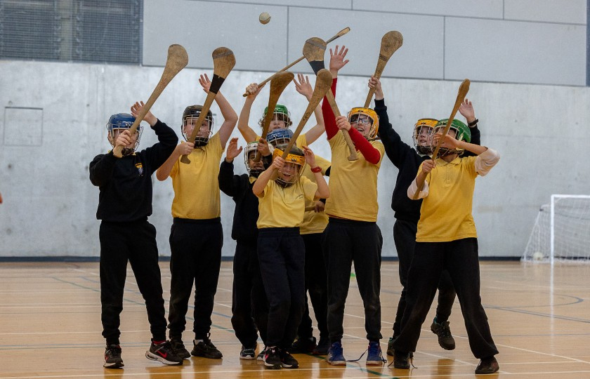 Seven primary school students wearing helmets and holding hurleys jostle playfully for a sliotar.