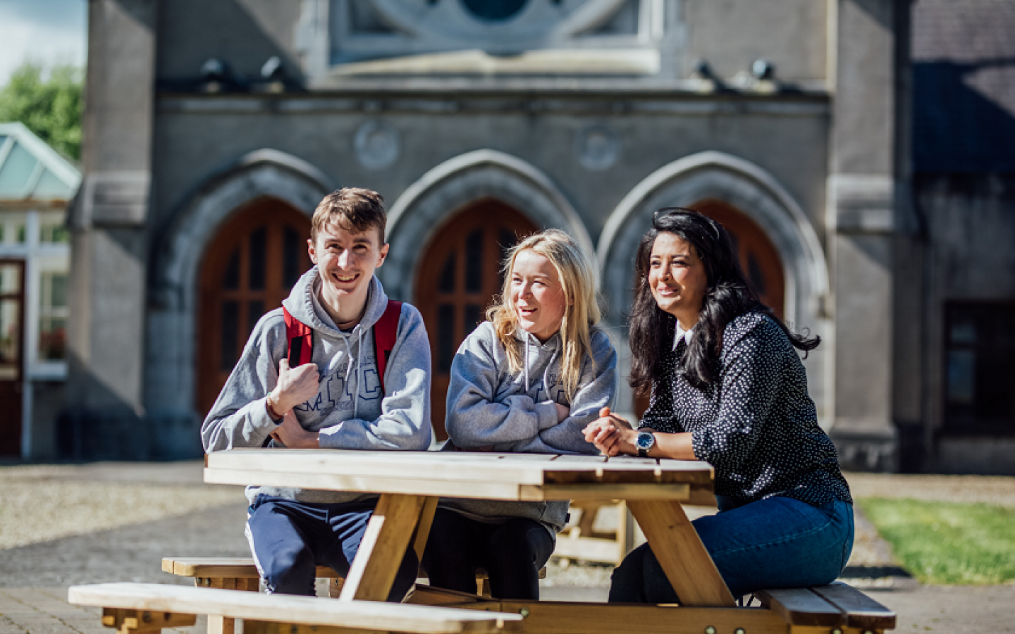 One male and two female students are pictured sitting at a wooden picnic table