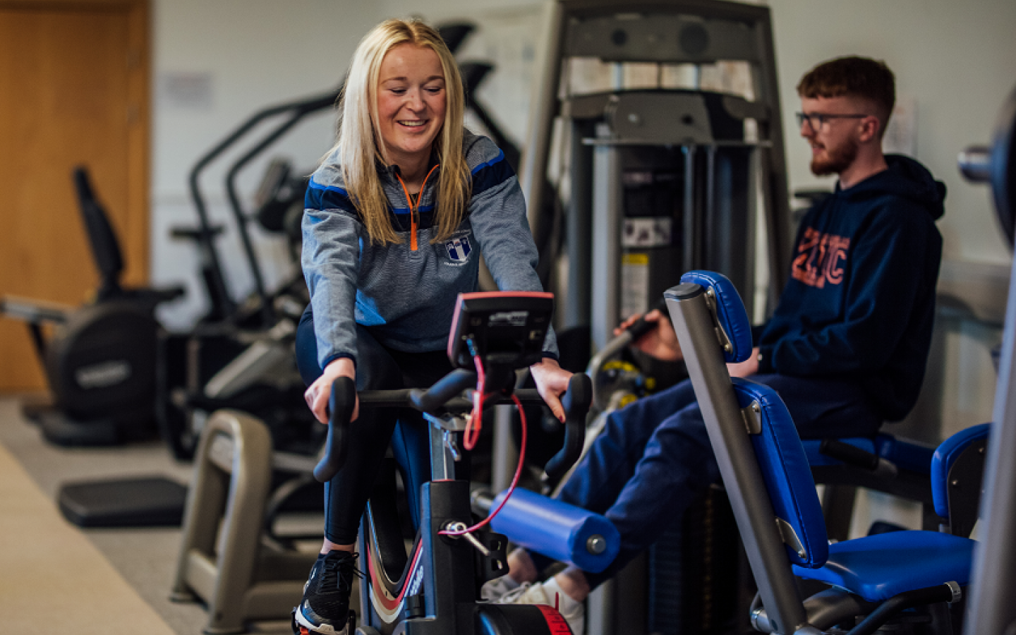 A female and male student are pictured in a gym using cycling equipment
