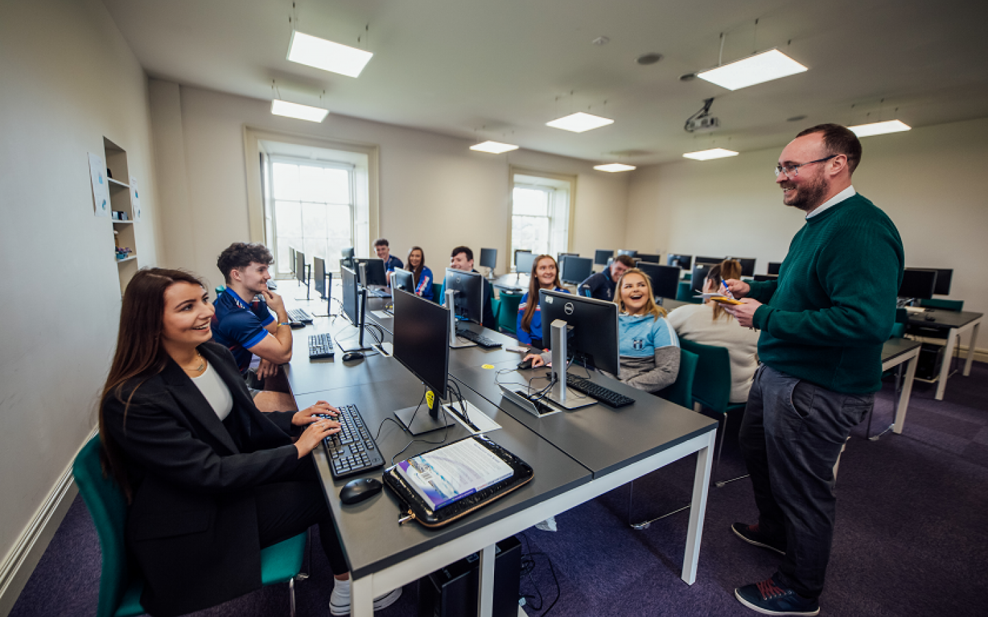 A group of students are pictured sitting at desks with desktop computer screens and keyboards.