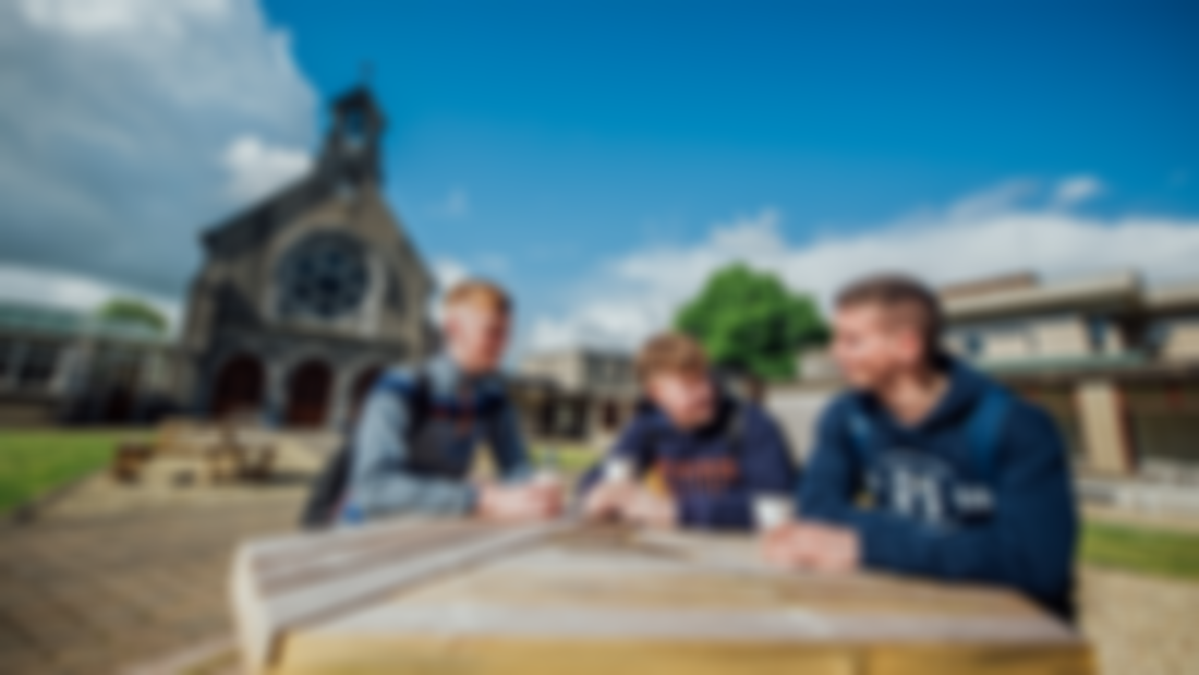 Three male students pictured sitting at a wooden picnic table. There is a chapel in the background.