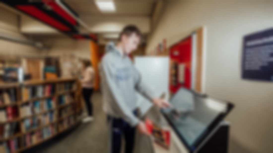 A male student is pictured in a library looking at a digital screen 