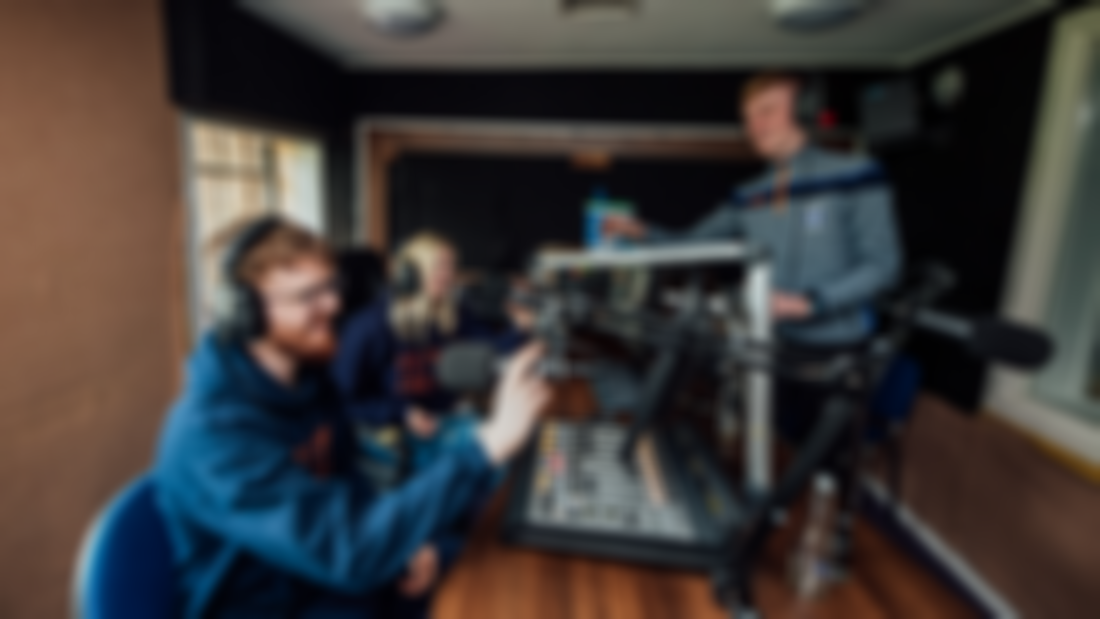 Two male and one female student are pictured in a radio studio with microphones and and audio mixing desk.