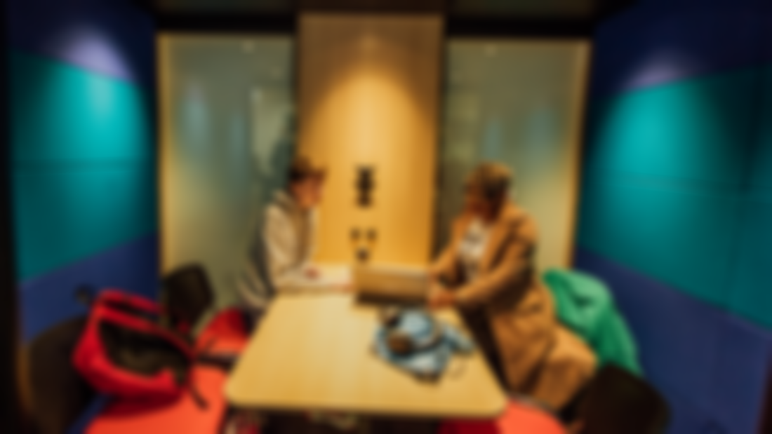 A male and female student are pictured in a study space with a desk and red chairs 