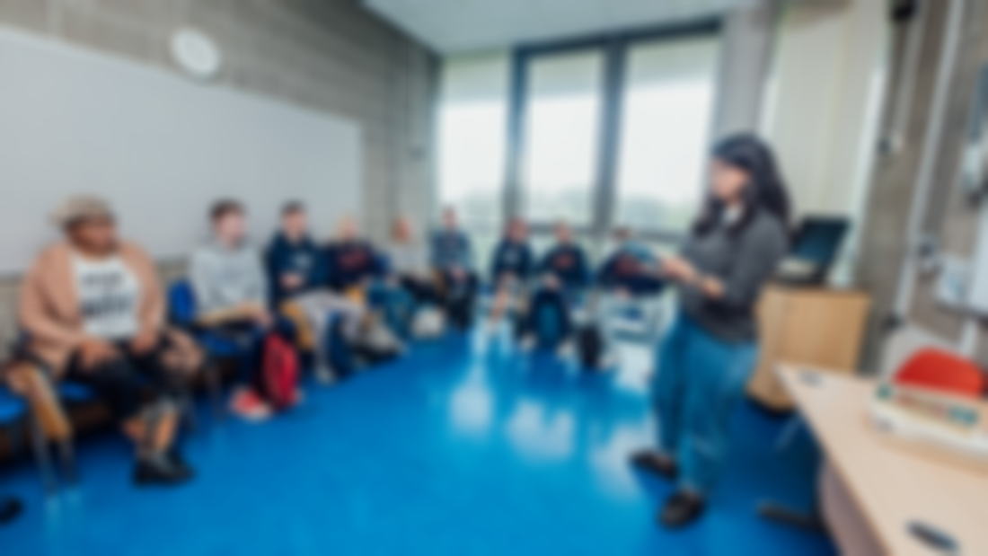 A group of students pictured sitting in a classroom with a female student standing reading from a book.