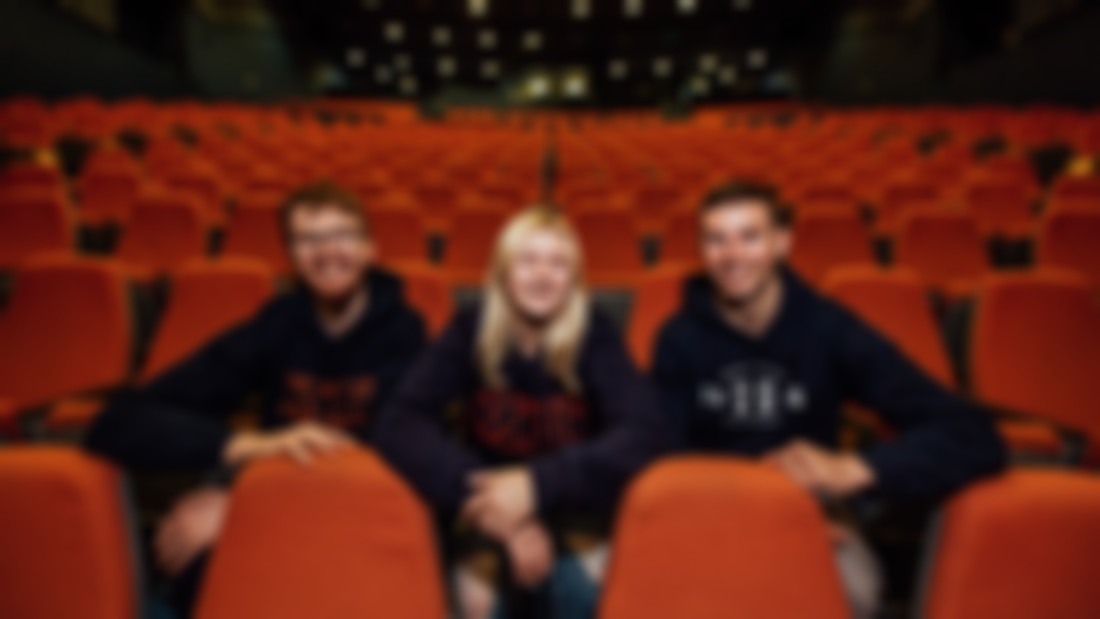 Two male students and one female student are pictured in a theatre with orange seats. 