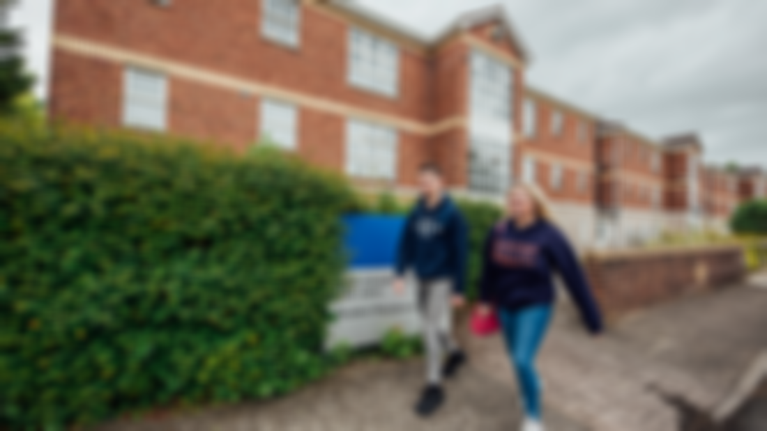 A male and female student walking on a footpath outside a large red brick building and green hedge.