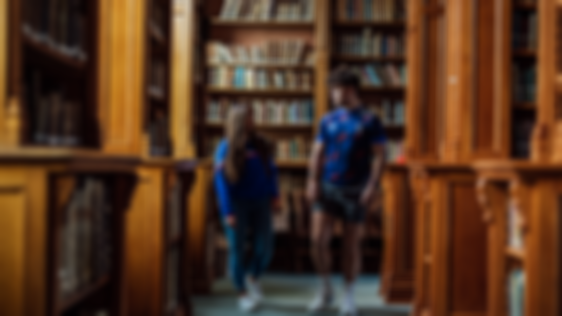 A female and male student are pictured walking through a library
