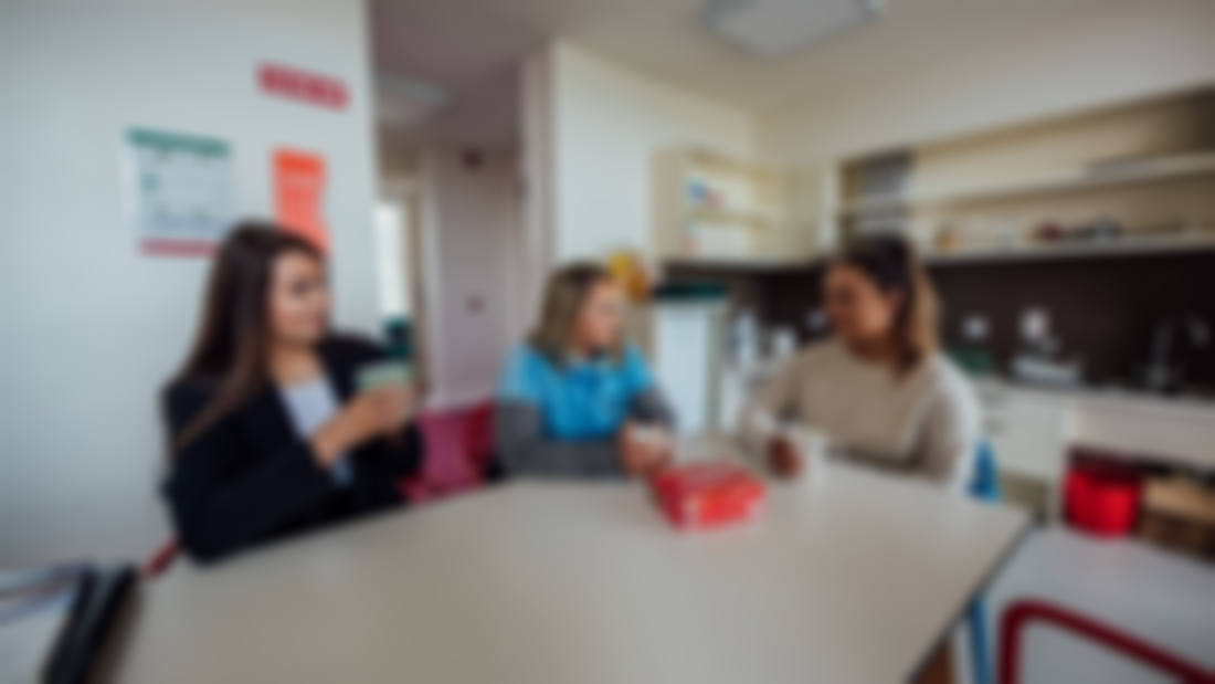 Three female students pictured sitting around a table in a kitchen chatting and drinking tea.