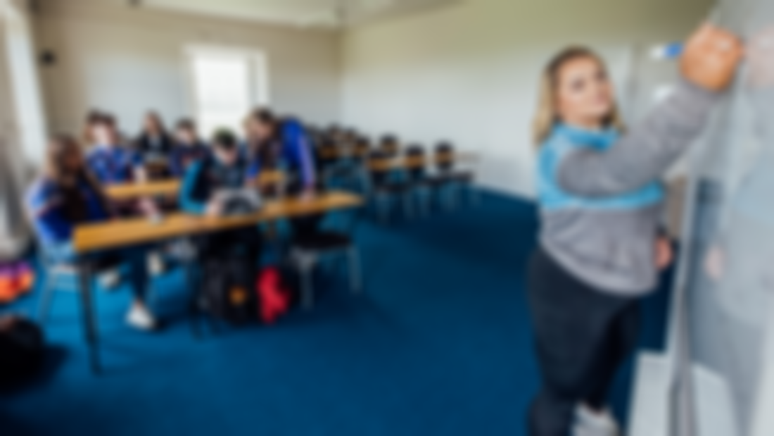 Students pictured sitting at desks in a classroom. A female student is pictured standing at the front of the classroom writing on a white board.