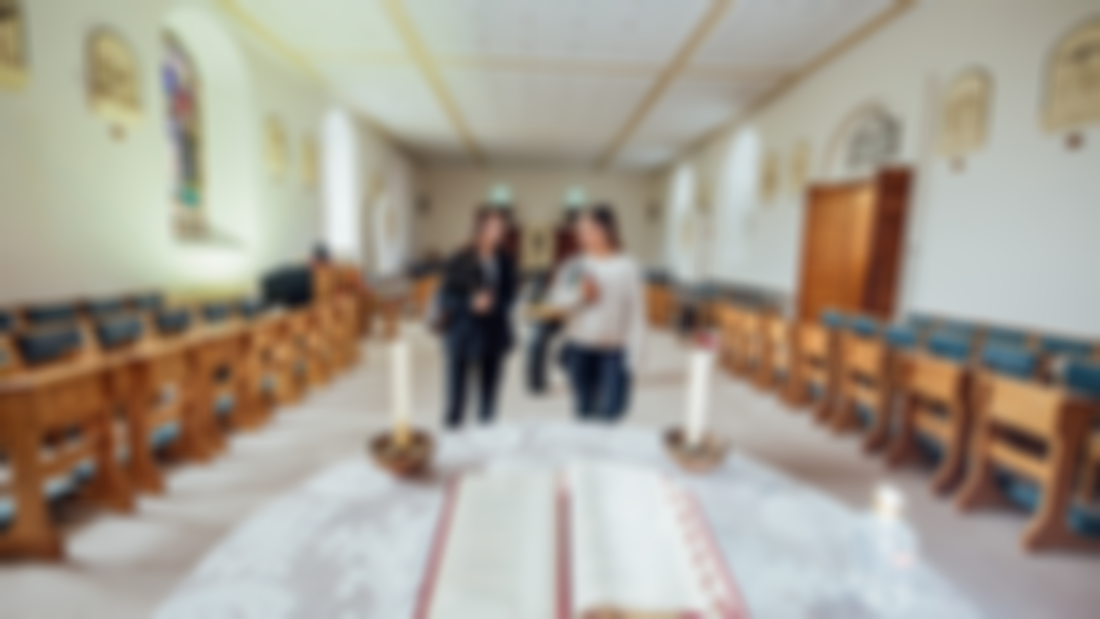 Two female students are pictured walking through a chapel with seats along either side and an alter with an open book in the foreground of the image. 