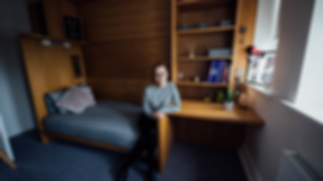 A female student pictured sitting on a bed in a bedroom with a desk and shelves.