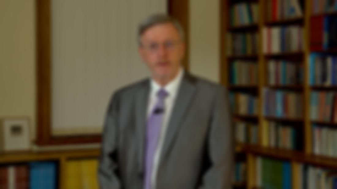 A male with short grey hair and glasses is pictured standing in a room with a shelf of books in the background. He is wearing a grey suit, white shirt and lilac tie.