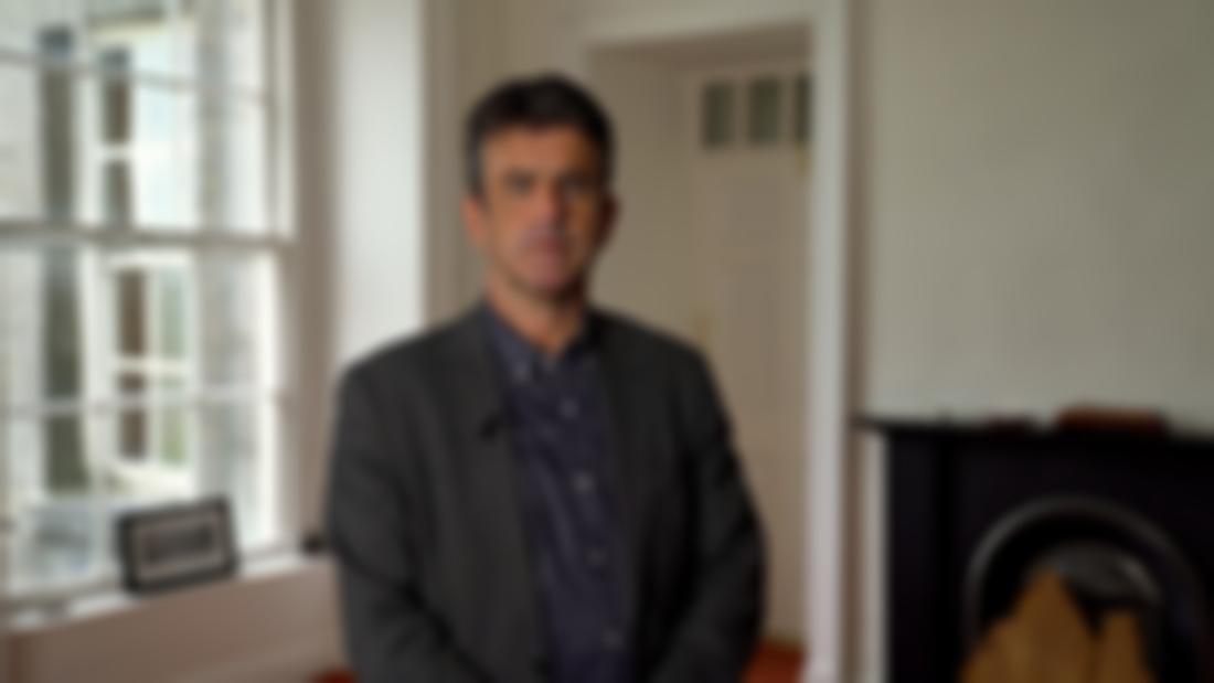 A male with short black hair stands in a room. He is wearing a blue shirt and grey blazer. 