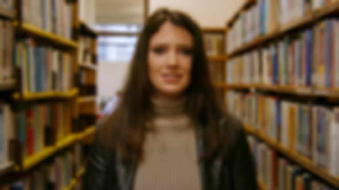 A woman wearing a leather jacket and beige polo neck. She is standing in a library with shelves filled with books