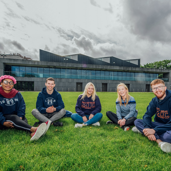 Three female students and two male students are pictured sitting on grass in front of a large modern building.