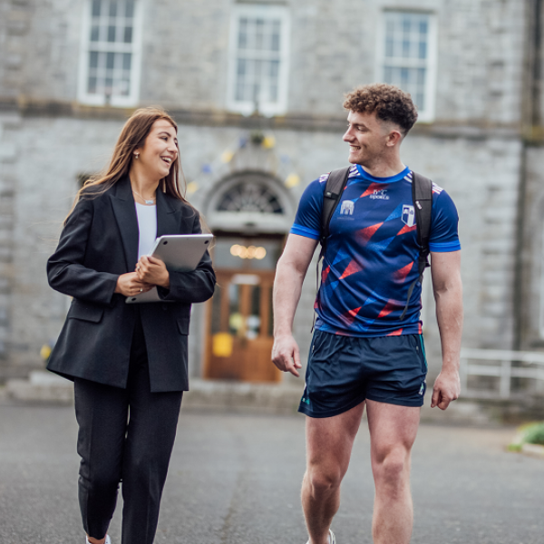 A female and male student are pictured walking and chatting to each other. In the background is a large 19th century building. 