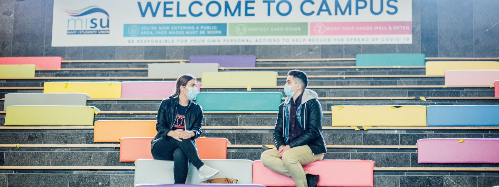 Two students with masks setting on TARA steps with Welcome to campus sign at their backs