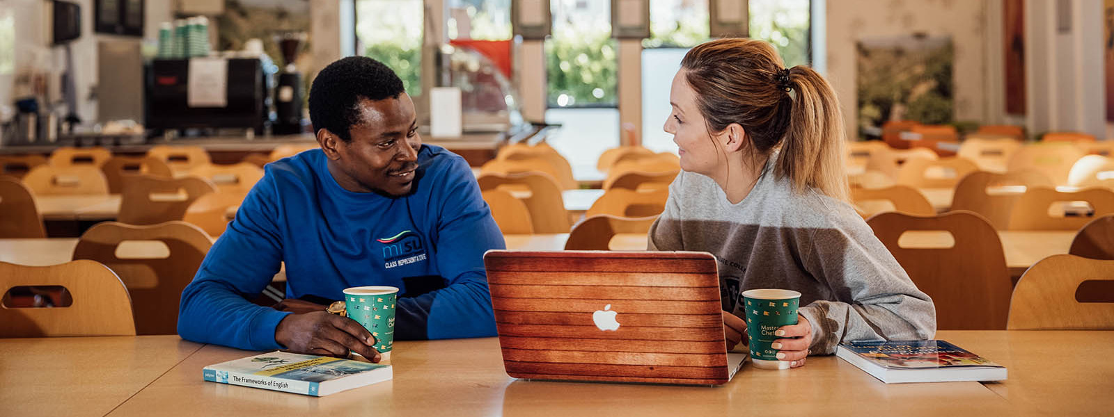 Two mature students sitting in the MIC canteen talking with a laptop open on the table.