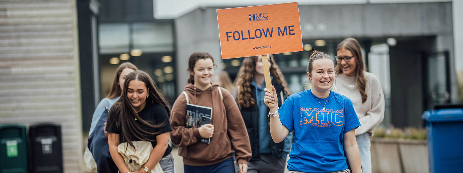 MIC student carrying a 'follow me' sign leading a group of visitors at a college open day.
