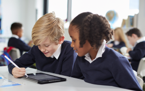 A male and female child dressed in school uniforms sitting a desk in a classroom and using an iPad