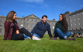 Two female and one male student sit on the grass in front of the MIC Thurles 19th century building 