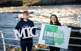 Two MIC students holding a Limerick.ie sign in front of the River Shannon
