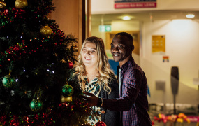 Male and female postgraduate students standing together looking at the Christmas tree in MIC Students Union.