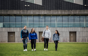 A group of four students are walking across a green field. There is a large grey building in the background.