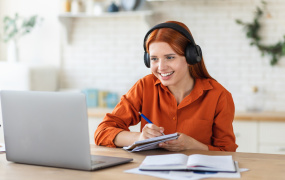 Female in orange top sitting at a table with a laptop and wearing headphones