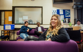 Female student with blonde hair sits on a purple couch looking over her shoulder with a smile at the viewer. There are a number of student sitting behind her in the background and are talking.