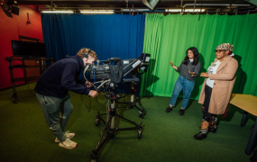 Male student behind a camera and two female students in front of camera in TV studio. There's blue and green screens set up behind them.