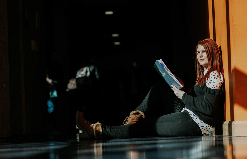 Woman sits on a floor holding a book and looking into the distance 