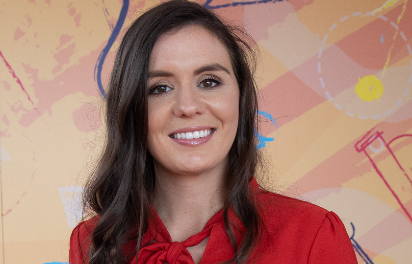 Presenter of Cúla4 Ar Scoil & MIC graduate, Orla Ní Fhinneadha, pictured in a red top against a beige background