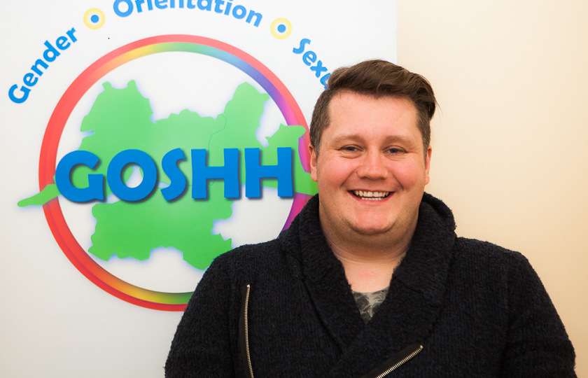 Patrick McElligott from GOSHH (Gender, Orientation, Sexual Health, HIV) standing in front of the GOSHH logo