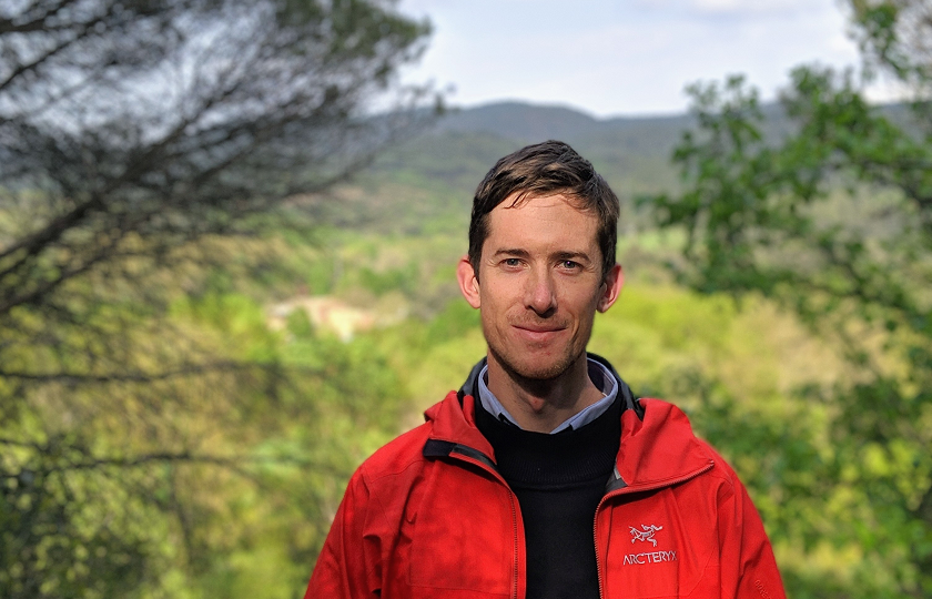 Dr Richard Butlet pictured wearing a red jacket and black jumper. He is standing outside with trees and fields in the background