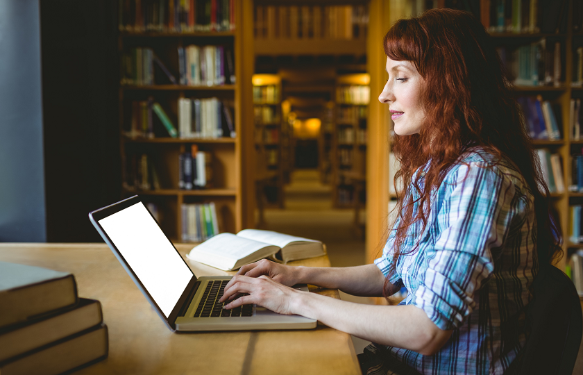 Side profile of a woman with red hair wearing a check shirt sits in a library typing on a laptop