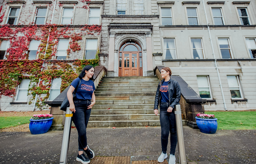 Two female students wearing MIC t-shirts and jeans lean on silver poles outside the steps leading up to a door on the Foundation Building at MIC