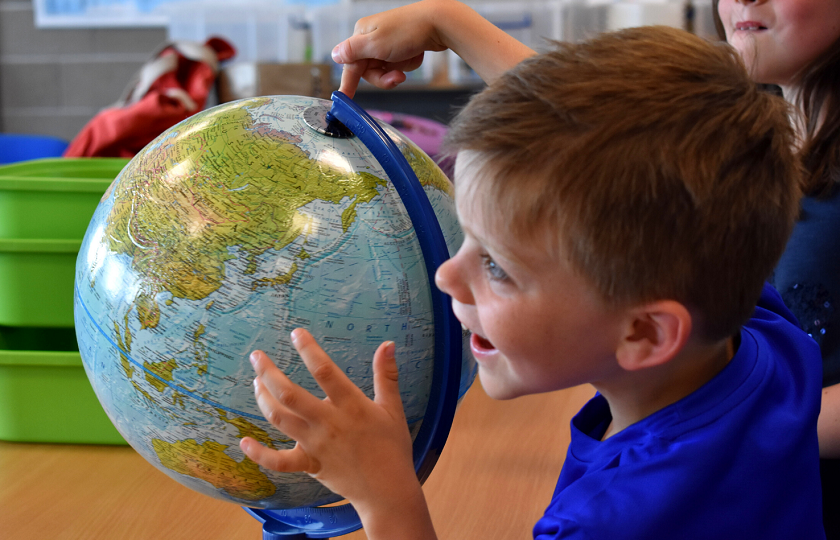 A male child wearing a blue t-shirt sitting in a classroom pointing at a globe 