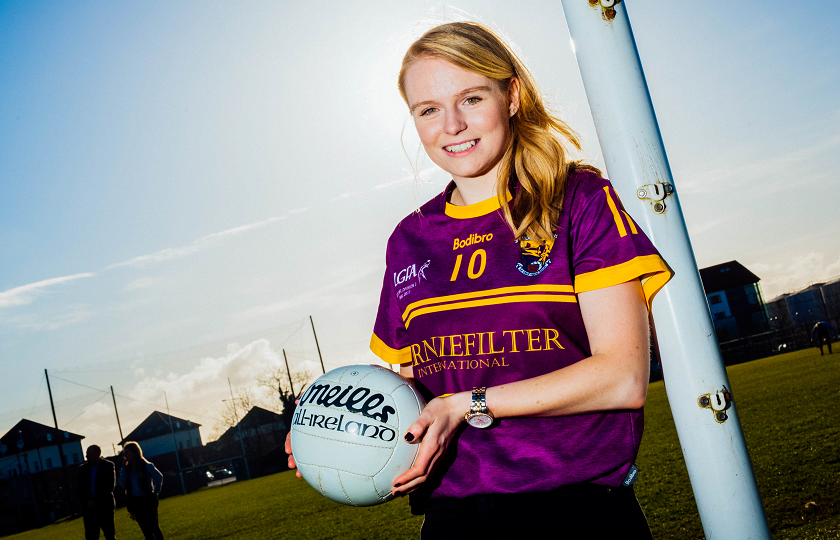 Female student holding a football and standing by a goalpost wearing a GAA jersey 
