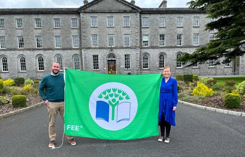 A man & woman pictured holding the Green Campus Flag on the grounds of MIC Thurles with the MIC Thurles building in the background