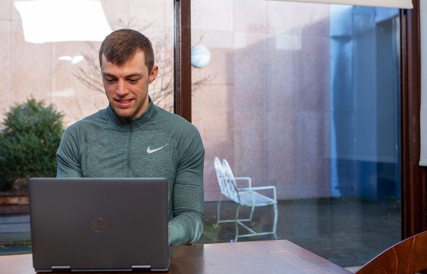 Male student wearing a green half zip Nike top smiles while looking at a laptop screen