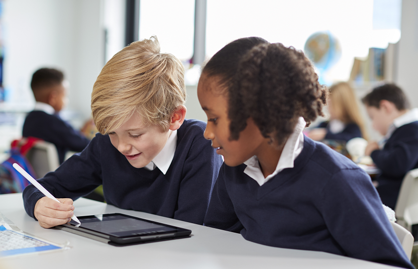 A male and female child dressed in school uniforms sitting a desk in a classroom and using an iPad