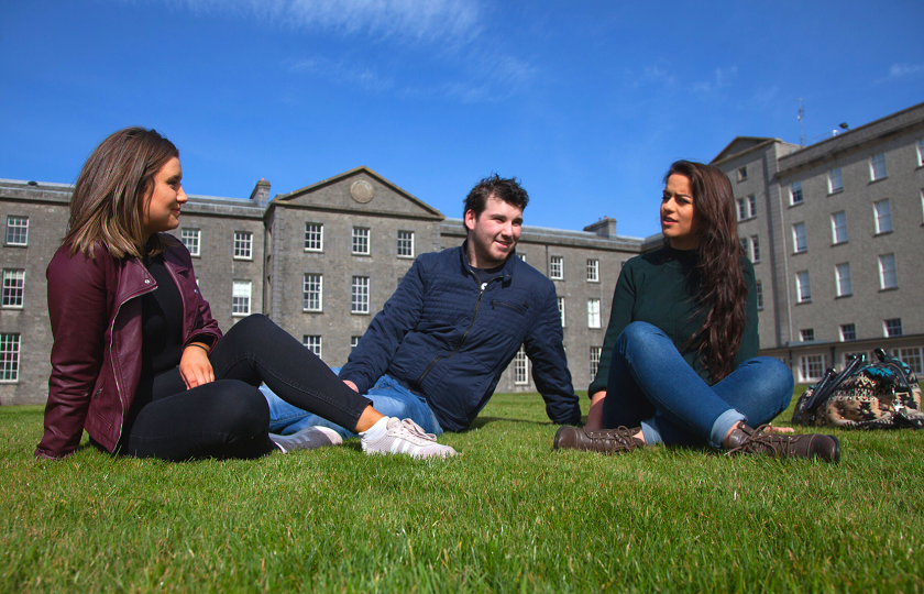 Two female and one male student sit on the grass in front of the MIC Thurles 19th century building 