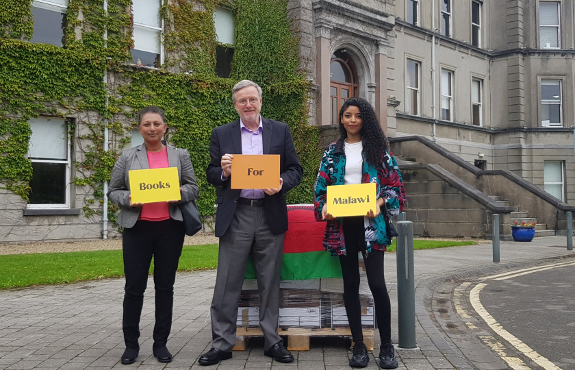 MC lecturer Santhi Corcoran, MIC President Prof. Eugene Wall and Mona-Lisa Das holding signs for Books for Malawi in front of a pallet of books
