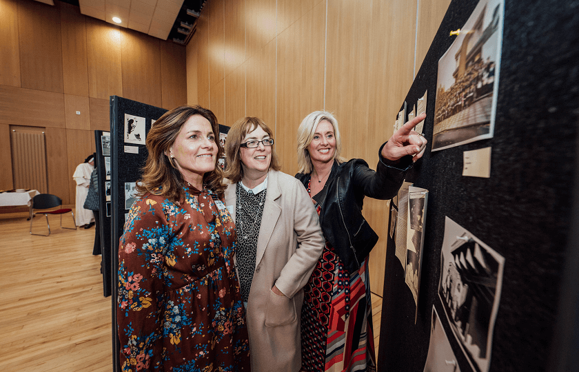 Three female alumni at a display of photos at the MIC Alumni Reunion 2019