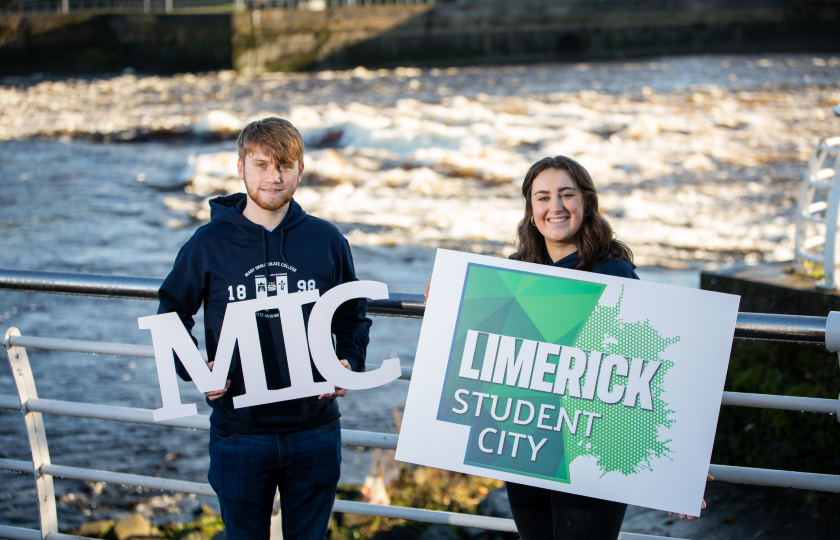 Two MIC students holding a Limerick.ie sign in front of the River Shannon