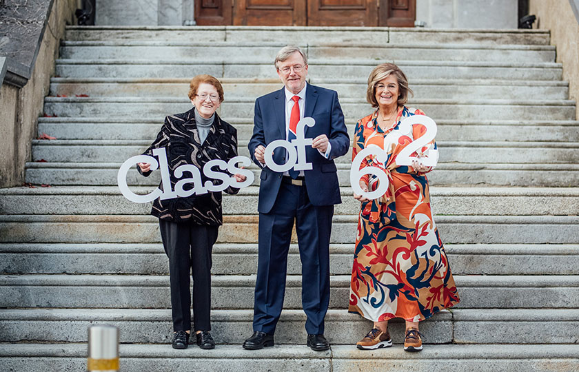 President Eugene Wall pictured with female alumna holding a sign Class of 62 on the steps of MIC foundation building.