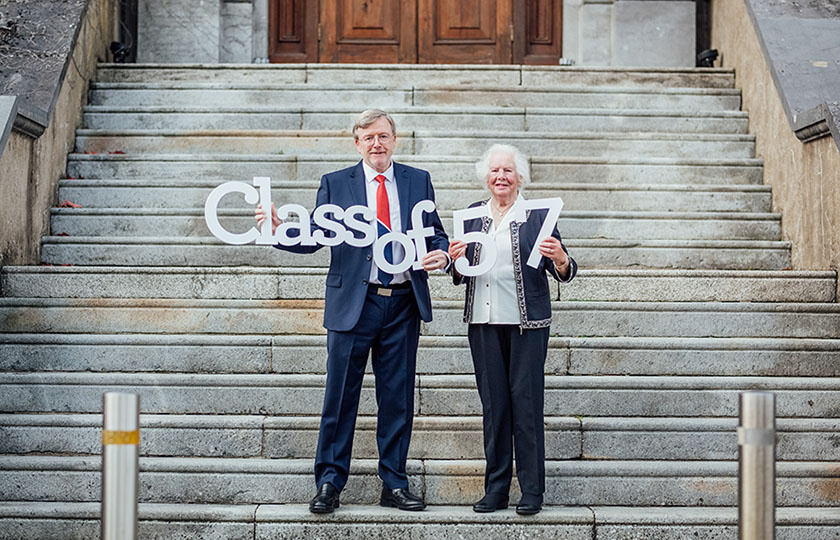 President Eugene Wall pictured with female alumna holding a sign Class of 57 on the steps of MIC foundation building.