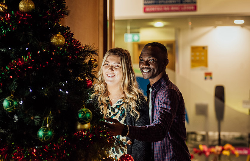Male and female postgraduate students standing together looking at the Christmas tree in MIC Students Union.